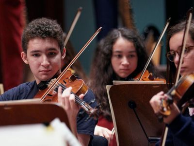 Three Junior Guildhall students on stage playing the violin concentrating and reading sheet music
